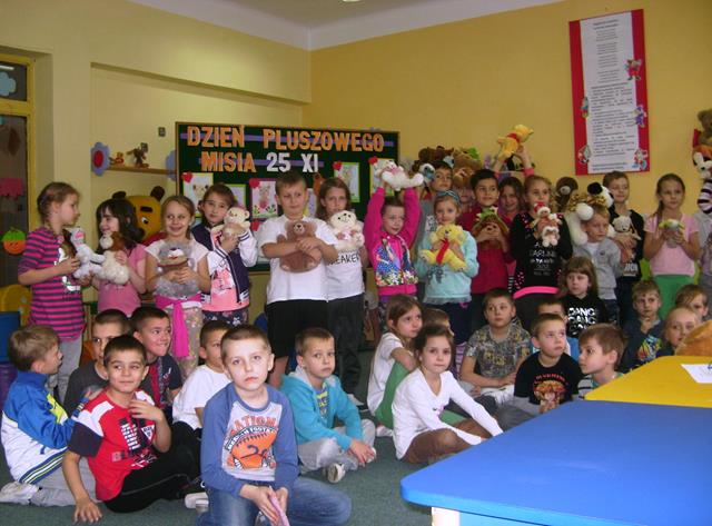 Zdjęcie przedstawia dużą grupę uczniów w świetlicy szkolnej. Dzieci trzymają w dłoniach pluszowe misie. W tle tablica z napisem: ,,Dzień Pluszowego Misia 25 XI’’.