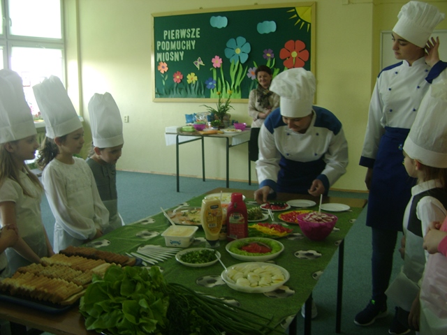 Zdjęcie przedstawiające dzieci podczas zajęć w świetlicy. Uczniowie stoją przy stole, na którym znajdują się różne produkty spożywcze. Dzieci przygotowują kanapki wspólnie z prowadzącymi zajęcia.
