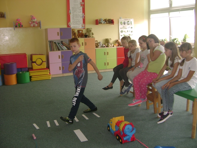 Zdjęcie przedstawia grupę uczniów w świetlicy szkolnej. Jedno z dzieci przechodzi przez symboliczne przejście dla pieszych zrobione z białych pasków papieru. Obok stoi plastikowy samochód. Pozostali uczniowie siedzą na stolikach.