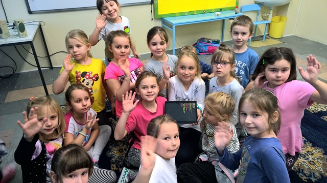Grupa dzieci z podniesioną ręką. Jedna dziewczynka trzyma tablet.