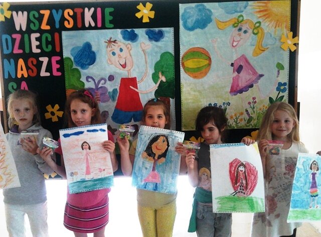 Zdjęcie przedstawia niewielką grupę uczniów. Prezentują prace plastyczne z wizerunkiem dzieci. W tle tablica z ilustracjami i  napisem “Wszystkie dzieci nasze są”.