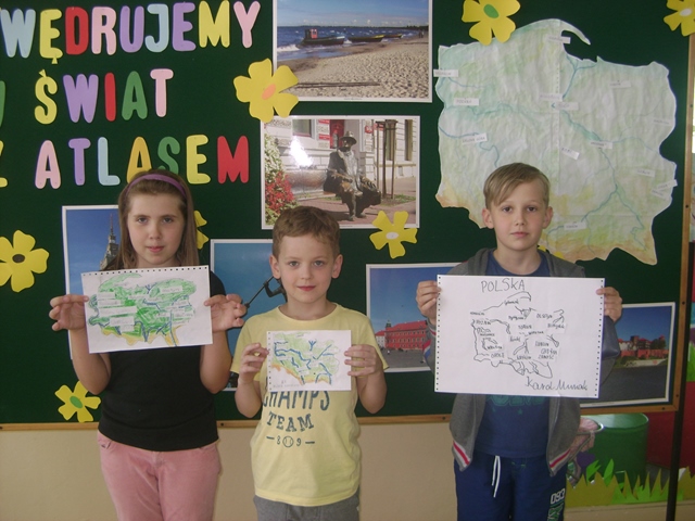 Zdjęcie przedstawia troje uczniów trzymających w rękach odręcznie wykonane mapy. W tle tablica z napisem “Wędrujemy w świat z atlasem” oraz zdjęcia z różnych regionów Polski.