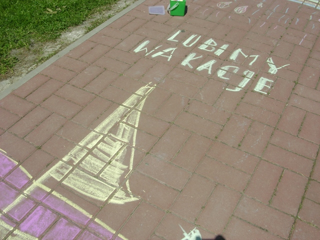 Zdjęcie przedstawia chodnik z rysunkiem łódki wykonanym za pomocą różowej i żółtej kredy. Wyżej znajduje się napis “Lubimy wakacje”.