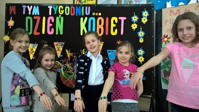 Grupa dziewczynek prezentuje na swych rękach bransoletki. Za nimi dekoracyjna tablica z napisem „W tym tygodniu... Dzień Kobiet”. Do tablicy przypięte są kolorowe kwiaty.