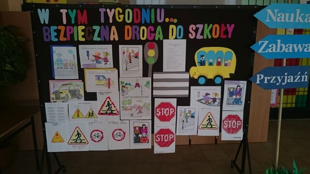 Dekoracyjna tablica z napisem „W tym tygodniu... bezpieczna droga do szkoły”. Pod hasłem przypięte są znaki drogowe i rysunki z różnych sytuacji na drodze.