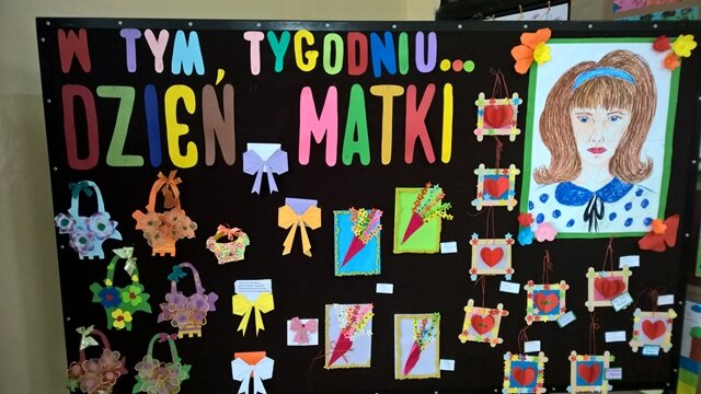 Dekoracyjna tablica z napisem „W tym tygodniu... Dzień Matki”. Do tablicy przypięte są ozdobne koszyczki, serduszka, bukiety oraz duży portret mamy.