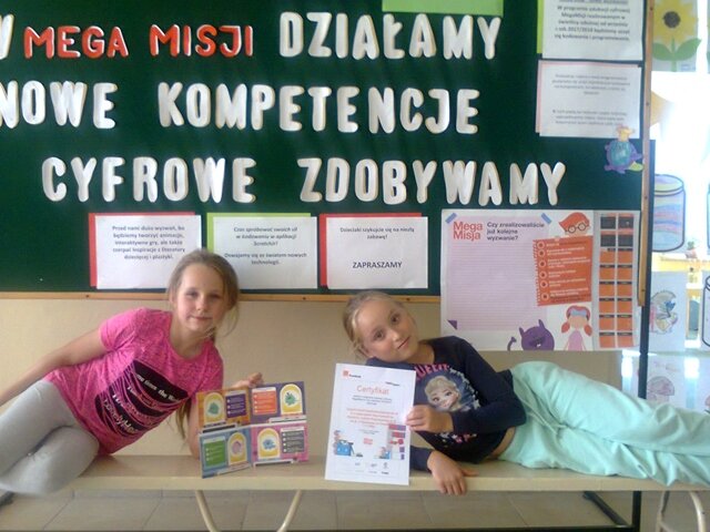 Dwie uczennice prezentują swoje dyplomy. Za nimi dekoracyjna tablica.