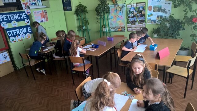 Uczniowie siedzą przy ławkach w grupach i wykonują zadania.