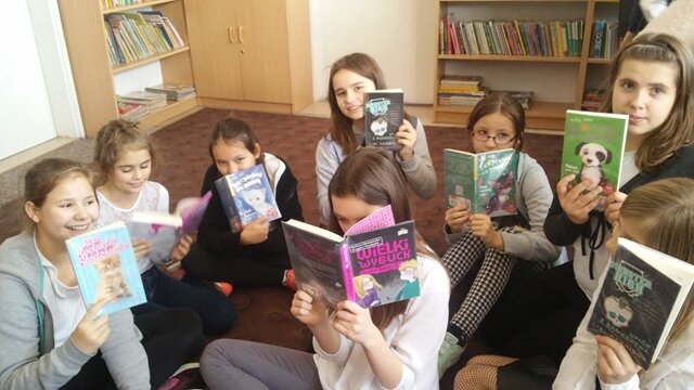 Uczniowie siedzą na dywanie w bibliotece szkolnej i trzymają w dłoniach książki.