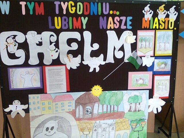 Dekoracyjna tablica z napisem „W tym tygodniu... lubimy nasze miasto” i rysunkami przedstawiającymi charakterystyczne atrakcje Chełma, m.in. Ducha Bielucha, studnię na rynku, niedźwiedzia między dębami.