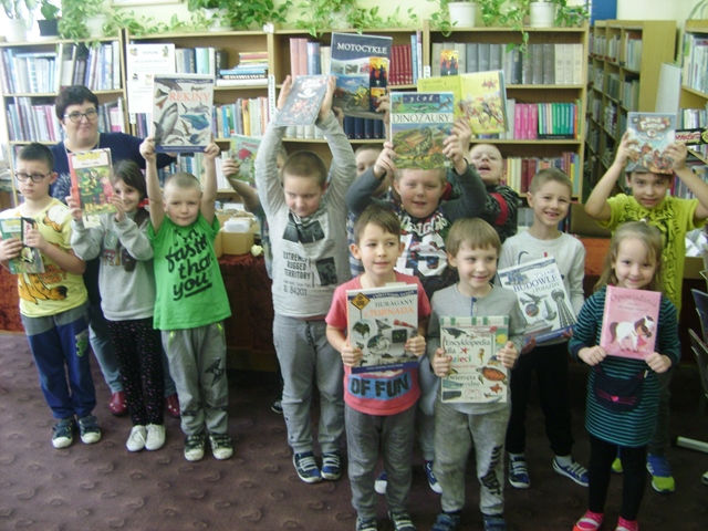 Zdjęcie przedstawiające uczniów i nauczycielkę w bibliotece szkolnej. Dzieci prezentują książki.