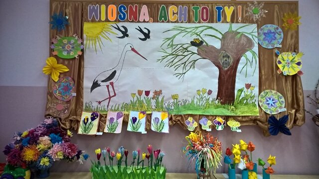 Gazetka ścienna o wiosennej tematyce. Na dużym rysunku przedstawiony jest bocian kroczący po łące kolorowych kwiatów. Nad nim świeci słońce, latają jaskółki, a on sam zmierza w stronę wysokiej wierzby.