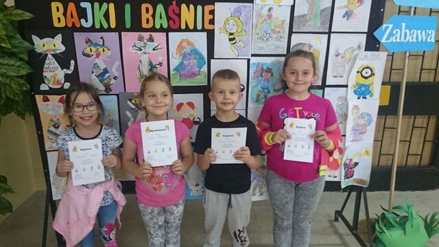 Czterech uczniów stoi na tle dekoracyjnej tablicy z napisem „Bajki i baśnie” z rysunkami bohaterów z bajek. Uczniowie trzymają w rękach dyplomy.