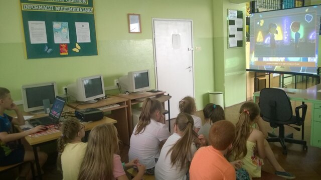 Grupa uczniów siedzi na ławeczkach i ogląda film na dużym ekranie.