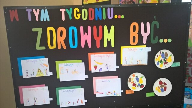 Dekoracyjna tablica z napisem „W tym tygodniu... zdrowym być”. Do tablicy przypięte są papierowe talerzyki z kolorowymi owocami.