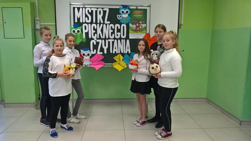 Uczennice prezentują maskotki serii "Gang  Słodziaków’’. W tle tablica z napisem "Mistrz pięknego czytania’’.