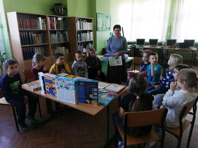 Zdjęcie przedstawia nauczycielkę i uczniów podczas zajęć w bibliotece szkolnej. Dzieci siedzą dookoła stołu, na którym znajdują się książki i gry planszowe. 