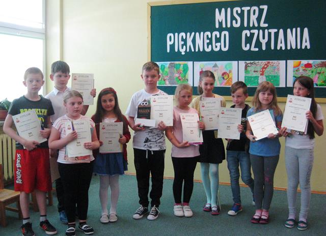 Uczniowie podczas rozstrzygnięcia konkursu pt. "Mistrz pięknego czytania’’. Dzieci stoją na tle udekorowanej tablicy i prezentują nagrody i dyplomy.