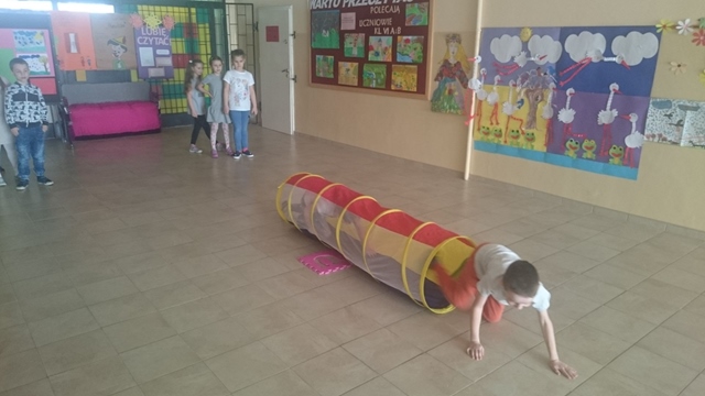 Uczniowie podczas zajęć sportowych. Dzieci wykonują ćwiczenia ruchowe, jeden z chłopów przechodzi przez tunel. 