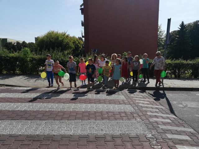 Grupa uczniów stoi przed przejściem dla pieszych. Uczniowie trzymają kolorowe balony.