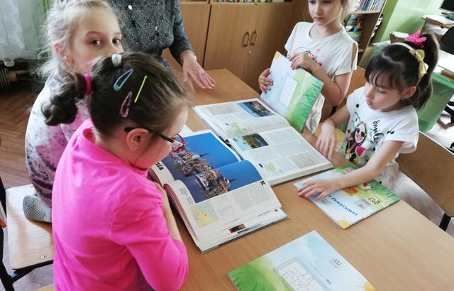 Uczennice klas młodszych siedzą przy ławce i czytają książki. 