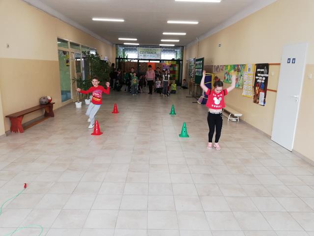 Dwie dziewczynki z podniesionymi rękoma biegną między pachołkami ustawionymi na korytarzu szkolnym, a w tle dzieci czekające na swoją kolej.