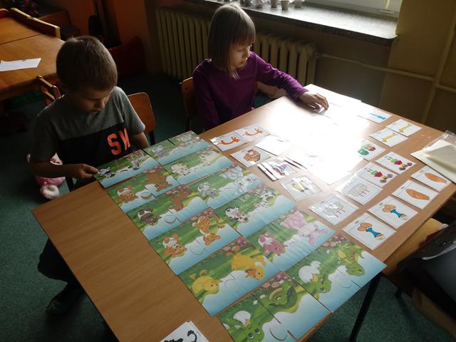 Dwoje uczniów siedzi przy stoliku, a przed nimi karty pracy z różnymi zadaniami.