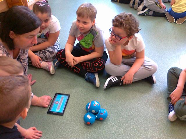 Uczniowie wraz z nauczycielką siedzą na dywanie wokół tabletu i robotów.