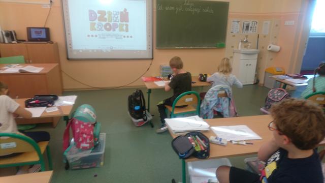 Zdjęcie przedstawia uczniów w sali lekcyjnej podczas zajęć. Na tablicy interaktywnej wyświetlony napis ,,Dzień kropki’’.