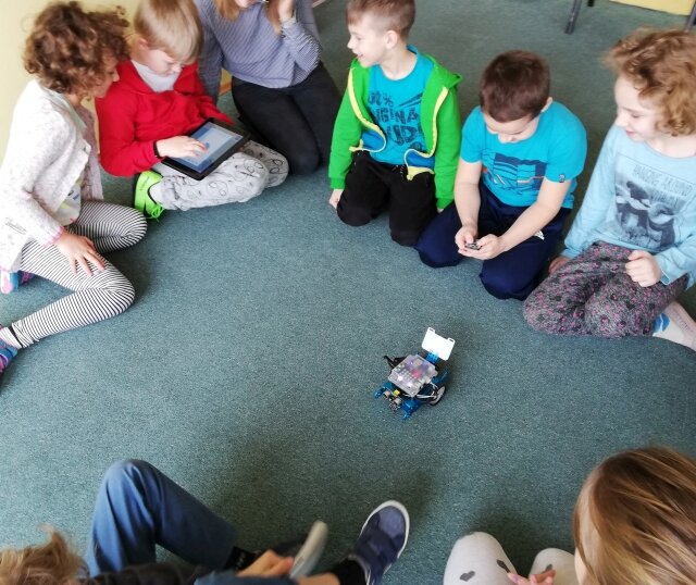 Zdjęcie dzieci, które siedzą na dywanie wokół małego robota.