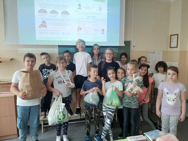Zdjęcie uczniów stojących pod tablicą interaktywną. Dzieci trzymają reklamówki pełne nakrętek.