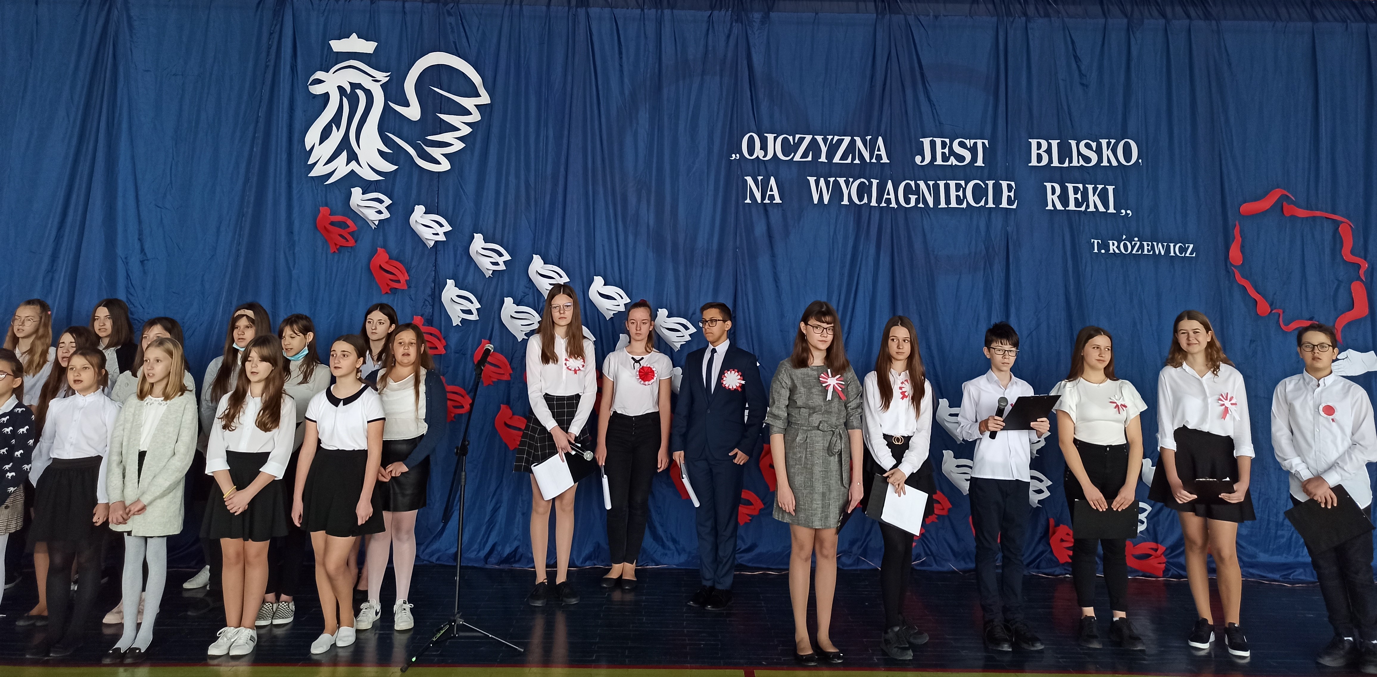 Zdjęcie grupy uczniów w strojach galowych, biorących udział w programie uroczystości obchodów rocznicy odzyskania niepodległości przez Polskę.