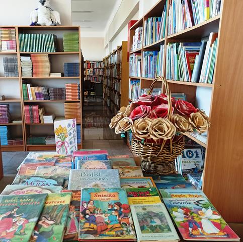 W szkolnej bibliotece na stoliku rozłożone są książki, a na nim stoi kosz z bukietem czerwono – złotych róż ręcznie wykonanych z suszonych liści. W tle znajdują się regały z książkami.