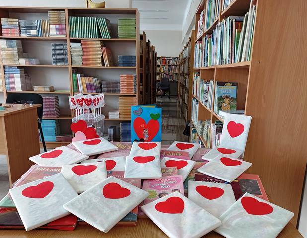 W szkolnej bibliotece na stoliku leżą książki, które są opakowane w biały papier z czerwonym sercem na środku. Obok nich stoją lizaki w kształcie serduszka. W tle znajdują się regały z książkami.