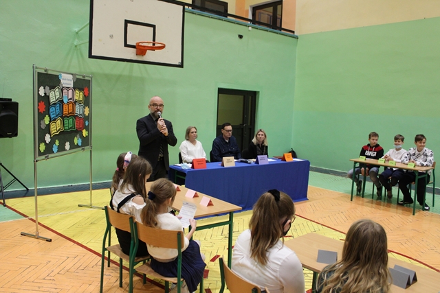 Zdjęcie przedstawia siedzących przy stołach uczniów i nauczycieli w sali gimnastycznej.