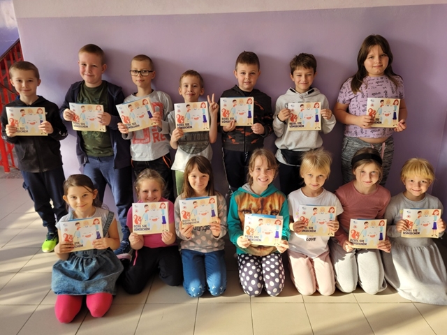 Zdjęcie grupy dzieci z pracami plastycznymi zachęcającymi do dzielenia się uśmiechem.