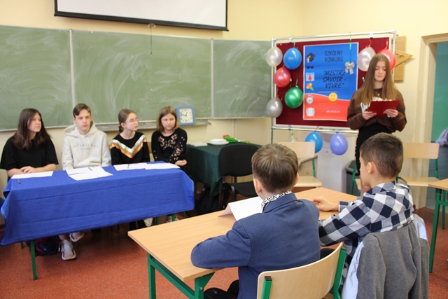 Zdjęcie przedstawia uczniów siedzących przy stolikach w sali lekcyjnej podczas konkursu.