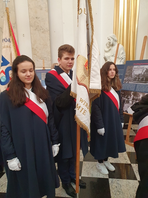 Zdjęcie pocztu sztandarowego - trojga uczniów w stroju galowym przepasanych szarfami biało-czerwonymi. Jeden z nich trzyma sztandar Szkoły Podstawowej nr 8 w Chełmie.