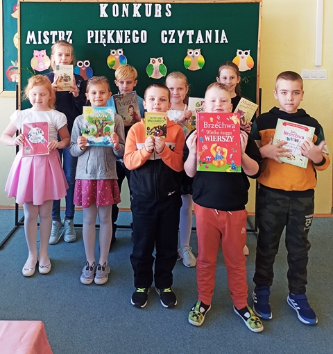 Na tle napisu; „Konkurs Mistrza Pięknego Czytania” grupa stojących dzieci prezentuje w dłoniach książki,  które są nagrodą w konkursie. 
