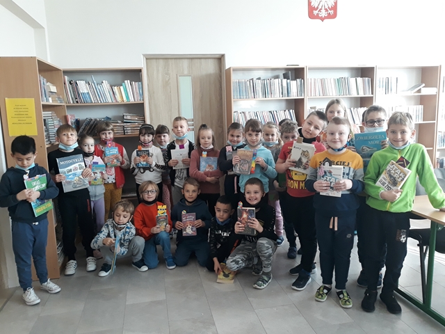 Zdjęcie przedstawia uczniów klasy pierwszej w bibliotece szkolnej. Uśmiechnięci uczniowie trzymają w rękach książki.