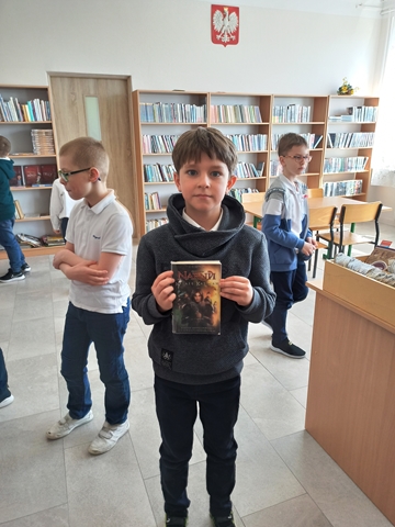 Zdjęcie przedstawiające uczniów podczas wypożyczania książek w bibliotece szkolnej. Na pierwszym planie uczeń prezentuje wybraną przez siebie książkę.