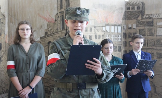 Zdjęcie przedstawia uczniów występujących w szkolnym przedstawieniu. Osoby zapowiadające ubrane są w stroje galowe, zaś uczniowie recytujący w szare mundury z biało-czerwoną opaską na ręku. 