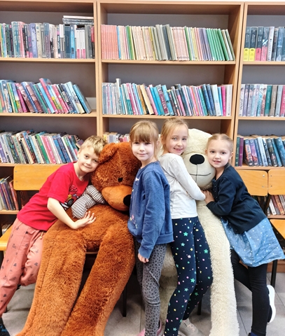 Zdjęcie przedstawia uczniów znajdujących się w bibliotece. Na fotografii widać trzy dziewczynki i jednego chłopca. Dzieci przytulają się do dużego białego oraz brązowego misia. 