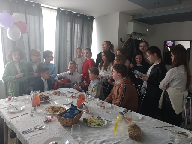 Zdjęcie przedstawia grupę uczniów zgromadzonych przy stole nakrytym białym obrusem, na którym znajdują się wigilijne potrawy i zastawa stołowa. Jeden z chłopców trzyma na kolanach gitarę.