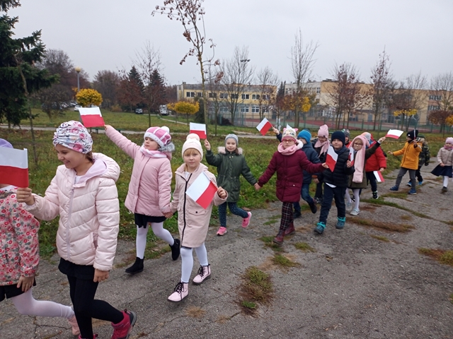 Zdjęcie przedstawia uczniów z klasy 1b uczestniczących w marszu niepodległości. Dzieci trzymają biało-czerwone flagi. 