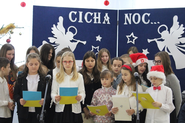 Zdjęcie przedstawia grupę uczniów podczas występu. W tle granatowa tablica z białymi aniołami i napis: “Cicha noc”.   