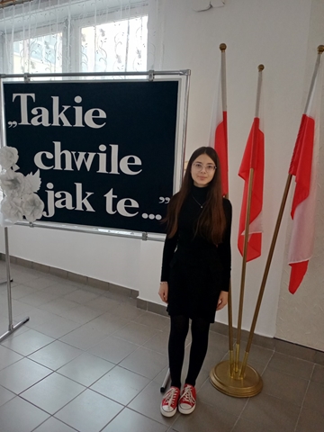 Uczennica stoi na tle polskich flag i tablicy z napisem:  „Takie chwile jak te…”.
