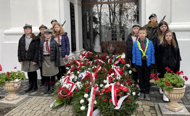 Na zdjęciu uczniowie w harcerskich mundurach stoją przy biało-czerwonych wieńcach podczas miejskich obchodów Święta Niepodległości. 