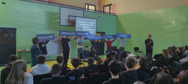 Zdjęcie ukazuje uczniów występujących na scenie oraz innych uczestników biorących udział w  zajęciach. Wydarzenie dotyczy programu rekomendowanego przez „Archipelag Skarbów”. 