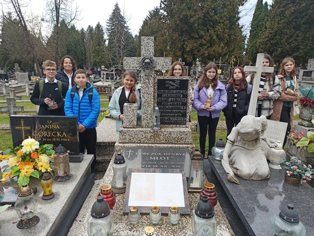 Zdjęcie przedstawia uczniów stojących za pomnikami. Dzieci ubrane są zimowe kurtki, a w dłoniach trzymają zapalone znicze. Na nagrobkach leżą sztuczne kwiaty i stoją znicze.